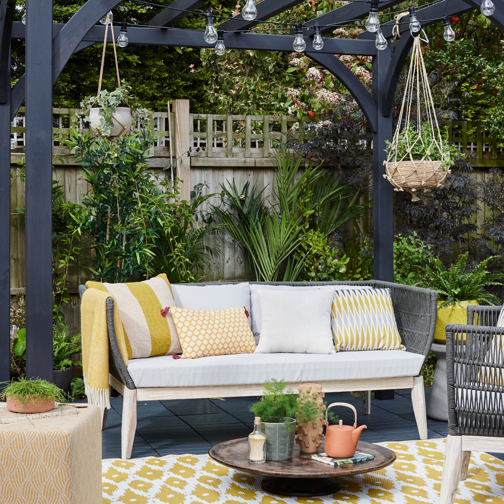 Garden sofa with white and yellow patterned cushions under a black pergola with plants in hanging baskets