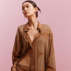 A model standing against a pink background wearing a loose brown shirt with white detailing done up by the central button.