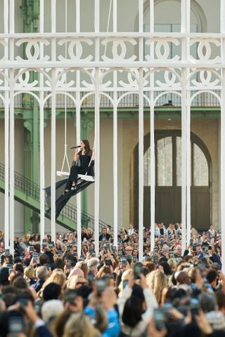 Riley Keough sitzt auf einer Schaukel im Grand Palais und trägt ganz schwarze Chanel bei der Modenschau Frühjahr/Sommer 2025, während er singt 