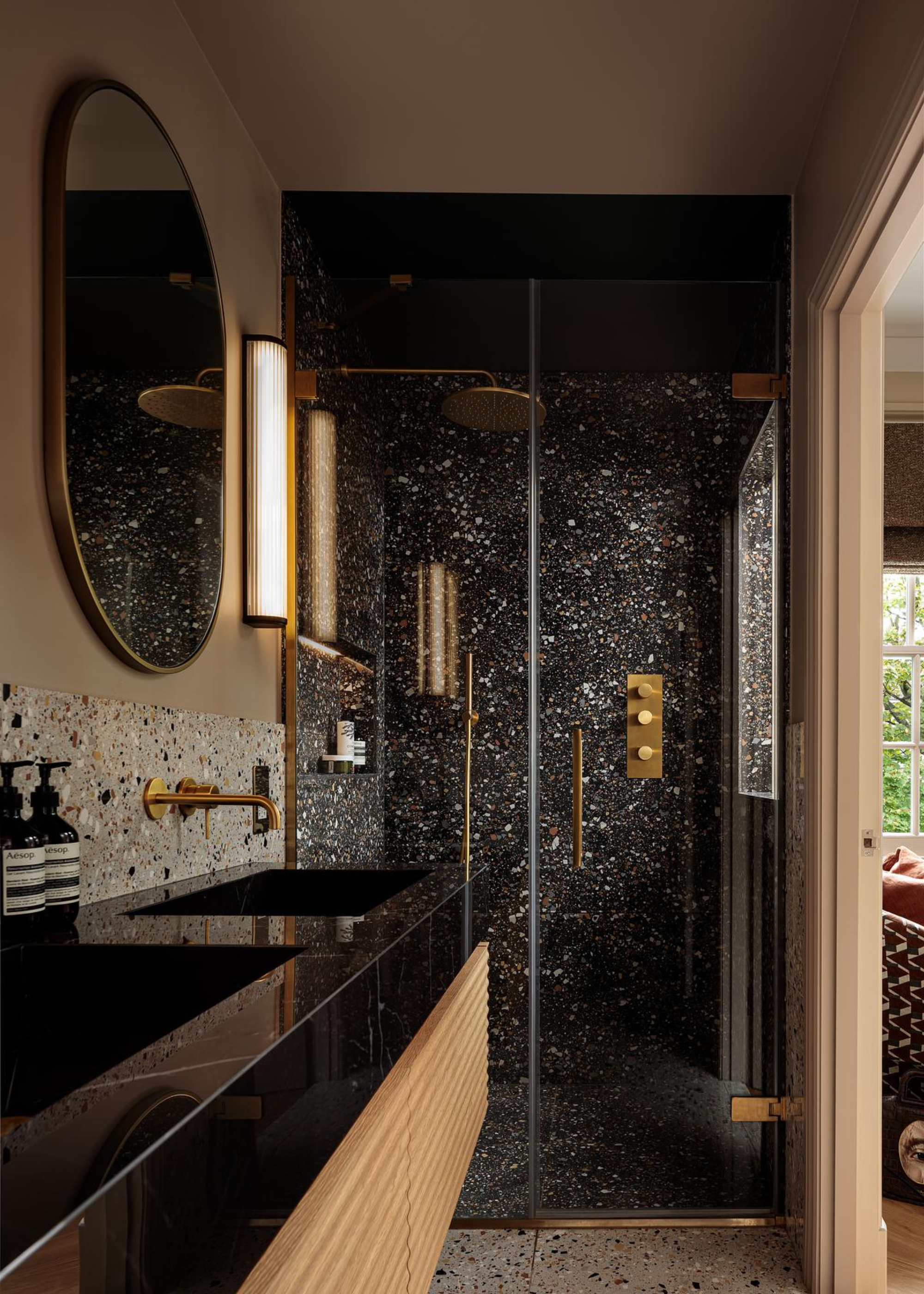 A deep and light terrazzo bathroom with a shower room, twin sinks, and Aesop toiletries near an elongated round mirror