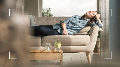 Woman smiling, lying down on sofa in living room wearing jeans and long t-shirt, a representation of why does heat make you tired