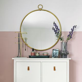 White chest of drawers against white wall with pink painted square, large round mirror on the wall above