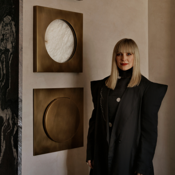 A headshot of interior designer Roisin Lafferty wearing a black pointed smart coat with her blonde hair worn down with a fridge and red lipstick standing in front of circular artwork