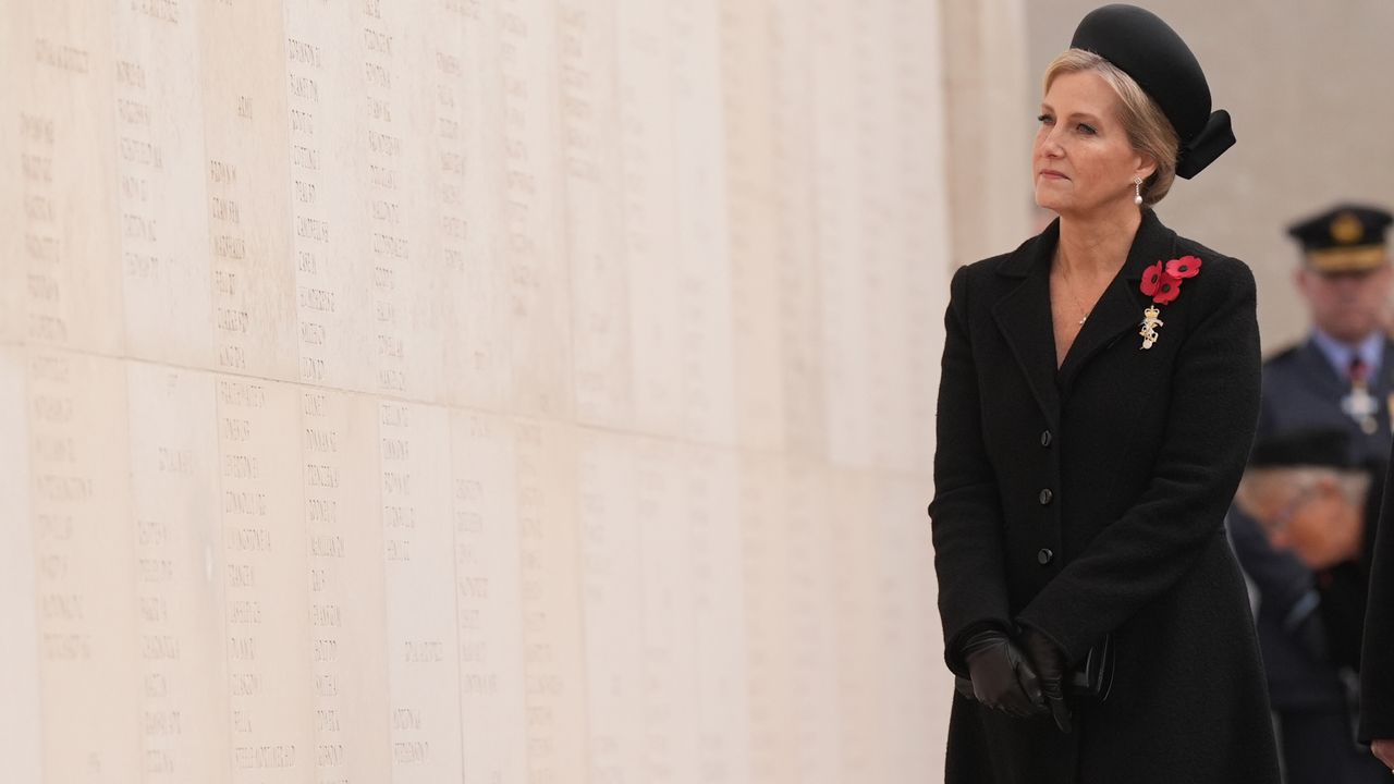 Duchess Sophie wearing a black coat and red poppy brooches standing in front of a war memorial wall