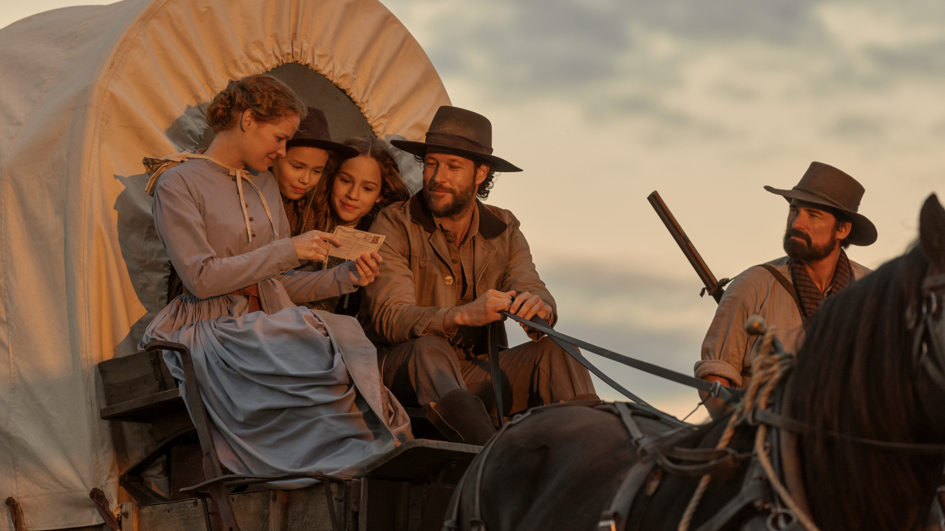 A still from Netflix's 'Little House on the Prairie.' A family (l-r Crosby Fitzgerald as Caroline, Alice Halsey as Laura Ingalls, Skywalker Hughes as Mary, Luke Bracey as Charles) sits on a covered wagon reading a letter as a man (Warren Christie as John Edwards) looks on from the right.