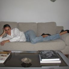 Anne-Laure Mais wearing a white button-down shirt, Levi's jeans, and ballet pumps while laying on a sofa inside her apartment. 