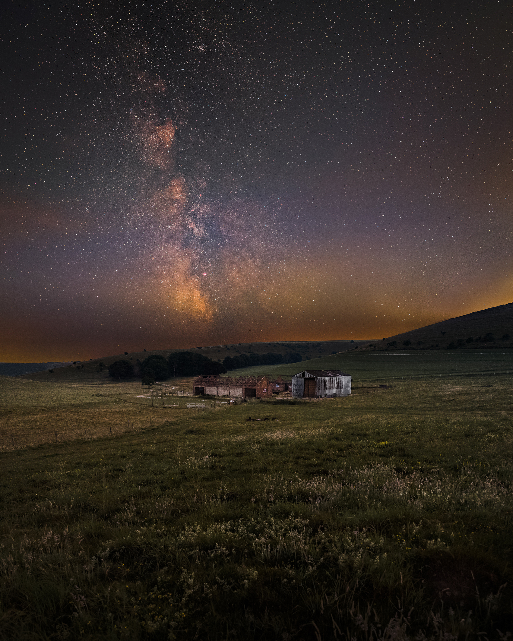 A serene rural landscape at night with an old barn on grassy fields under a starry sky. The Milky Way glows brightly, enhancing the tranquil mood