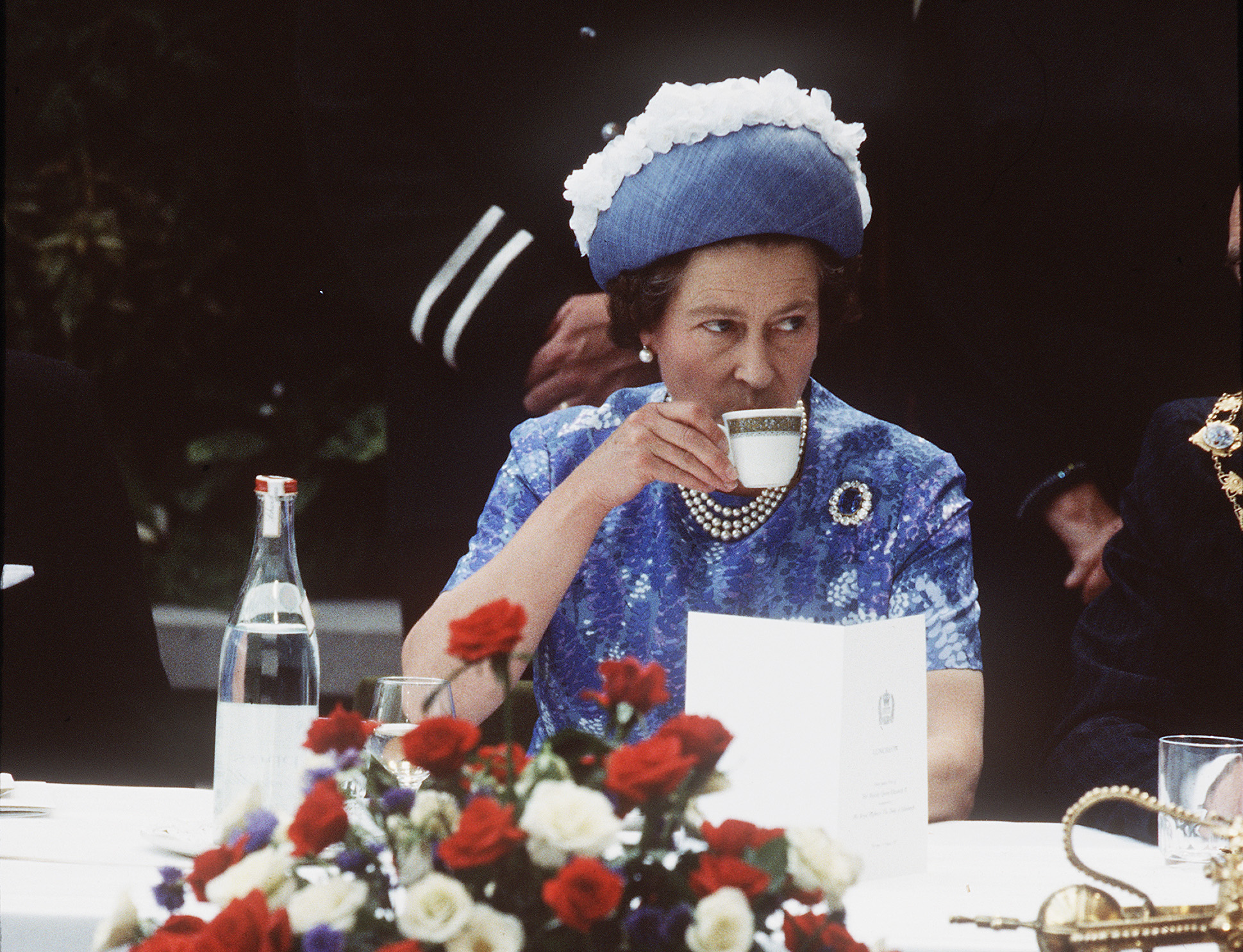 Queen Elizabeth wearing a blue dress and hat drinking tea at a table