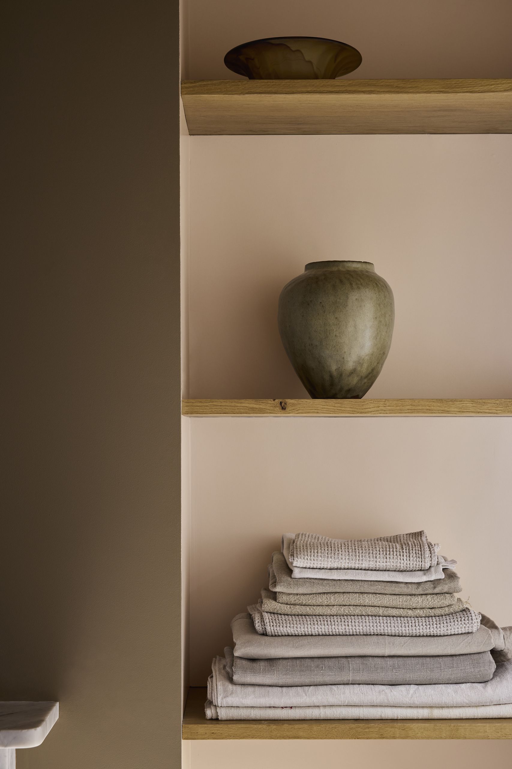 A bathroom shelf with brown walls and light pink at the back of the shelving. There is a stack of neutral towels and a ceramic vase.