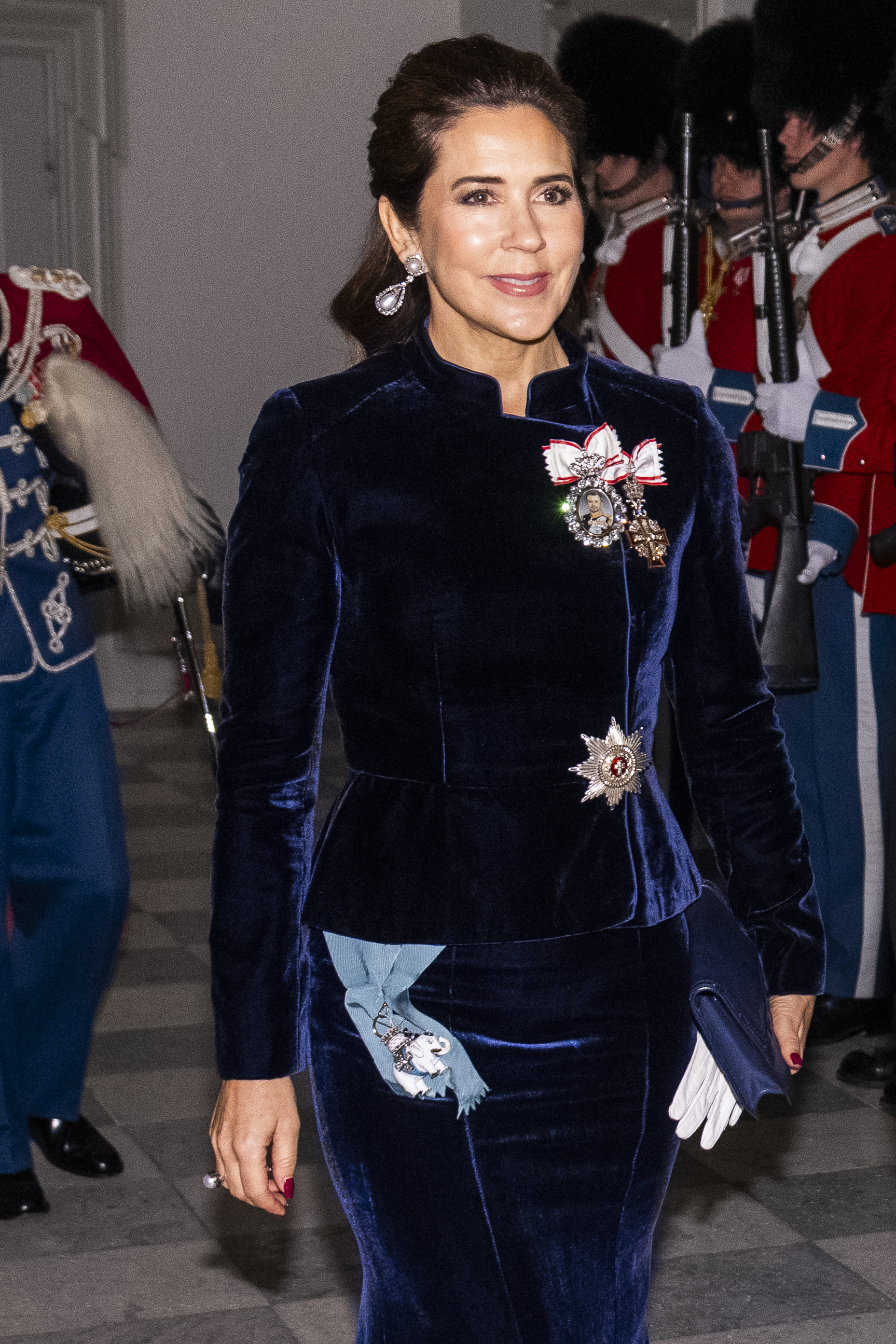 Queen Mary wearing a navy velvet dress and dangling earrings walking past royal guards