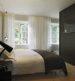 Hotel Skeppsholmen, Stockholm. A hotel room with a white bed, a black side table, wall lamps, a large dark wood cabinet and two windows with white curtains.