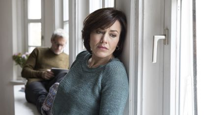 A mature woman looks unhappy as she looks out the window while an older man reads behind her.