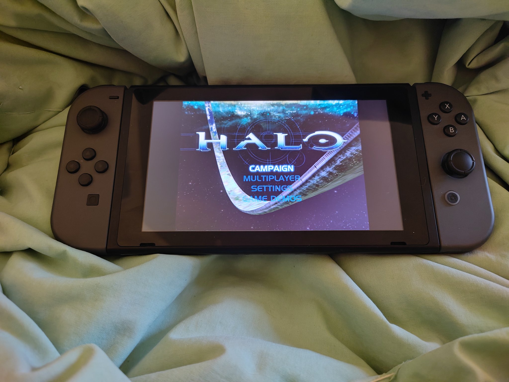 A Nintendo Switch displays the Halo title screen, showcasing options like Campaign and Multiplayer. It's resting on a crumpled, soft green blanket.