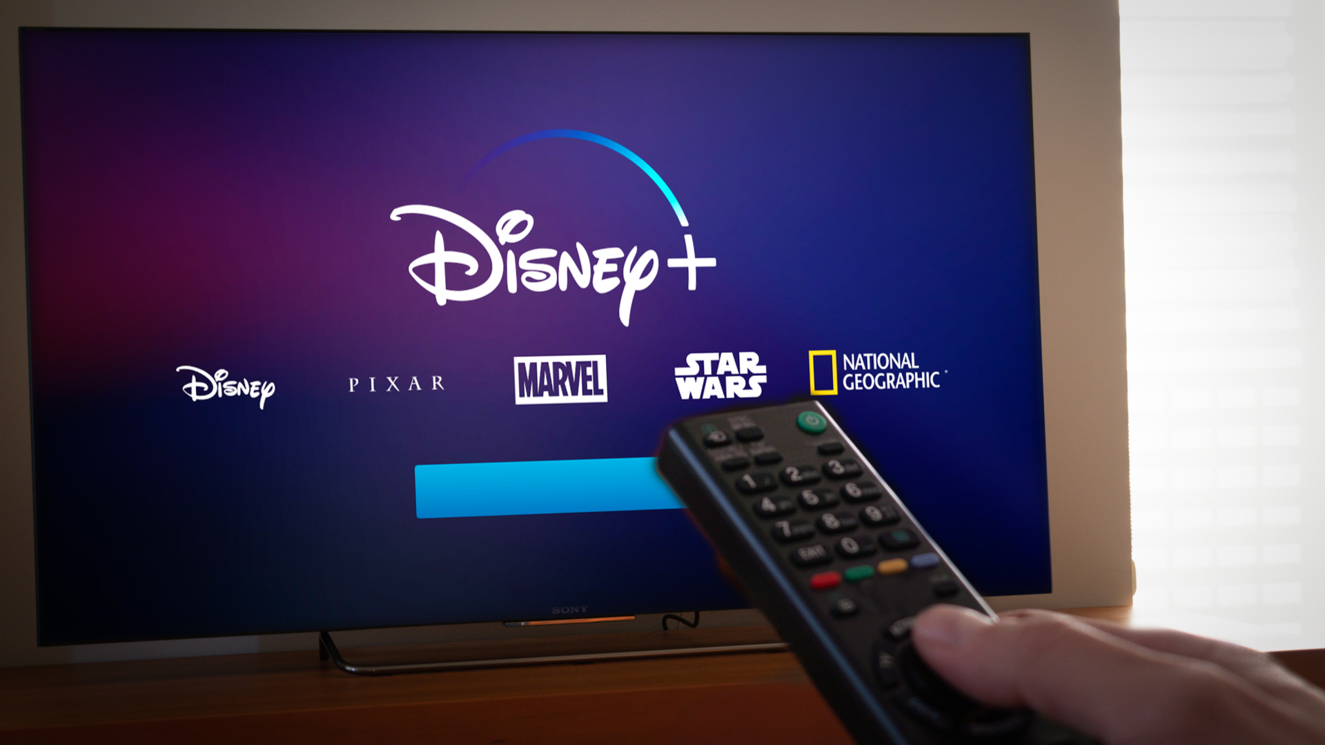 Un hombre sostiene un control remoto hacia una pantalla que muestra el logotipo de Disney Plus.