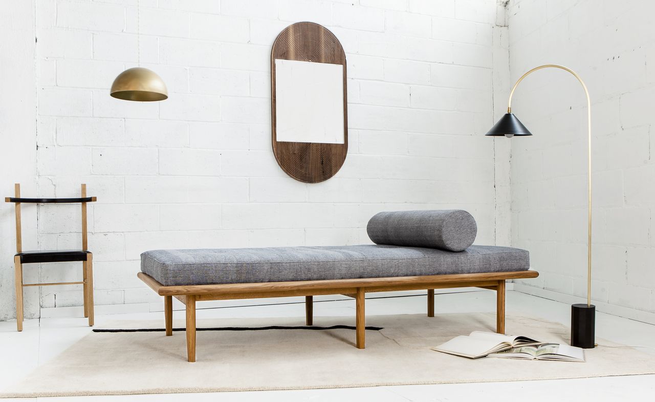  Room with white brick walls and floors featuring a capsule shaped mirror embedded in brown wood hanging vertically on the wall; ; wooden chair with black leather trims against the white brick wall; Floor lamp with black head and base connected by gold metal stand next to a grey day bed with brown wood base