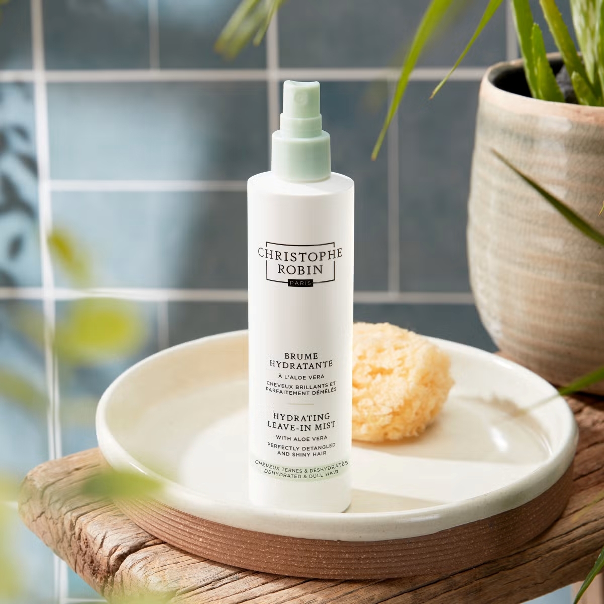Christophe Robin hydrating leave-in mist on bathroom stool with blue tiles and plants.