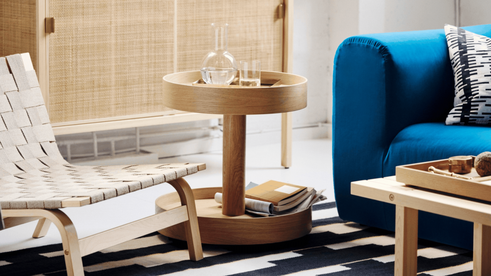 STOCKHOLM 2025 Side Table on Casters in Oak Veneer between blue sofa and woven armchair, on black-and-white patterned rug, in front of a woven rattan cabinet