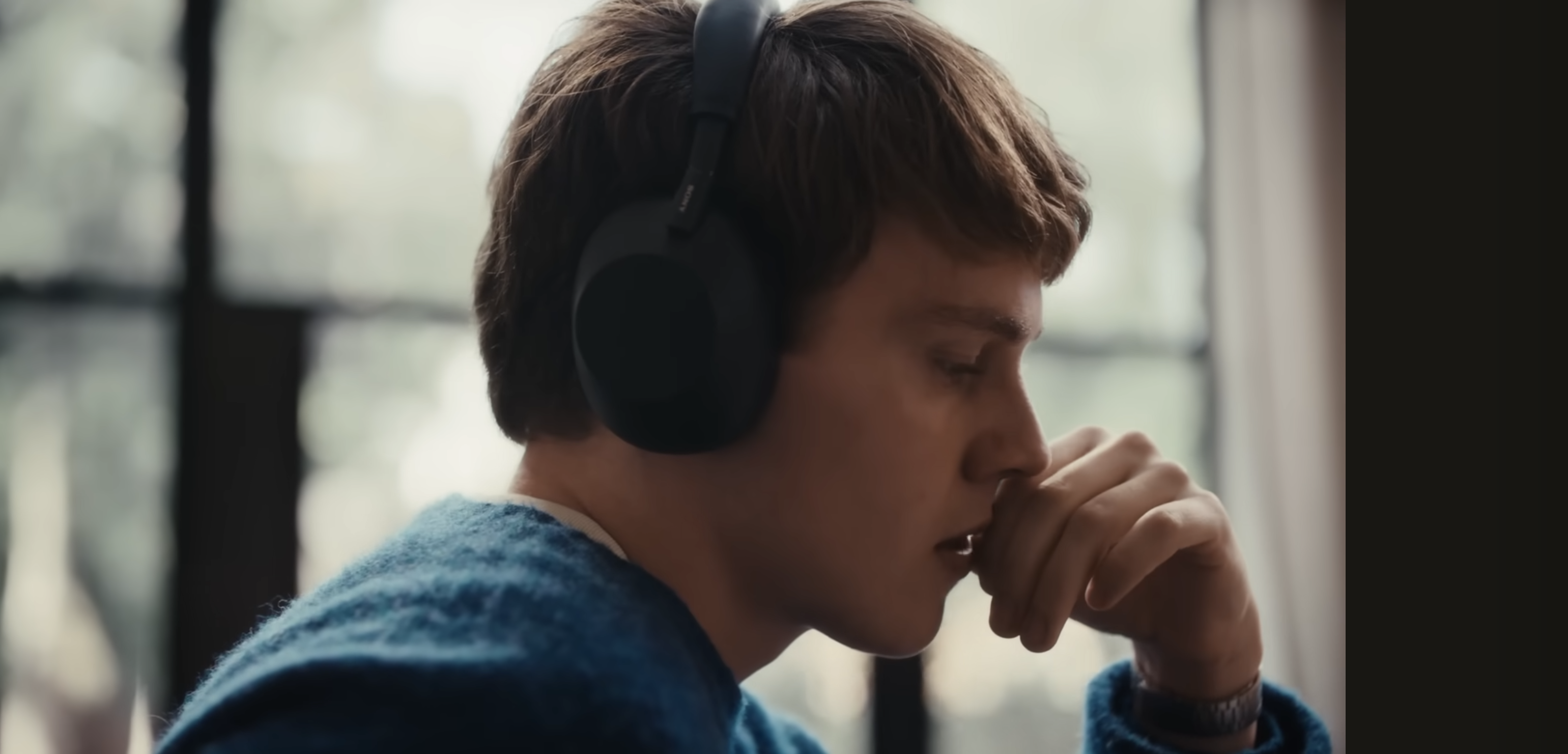 A young boy wearing a pair of blue headphones and blue jumper holding his hand up against his mouth