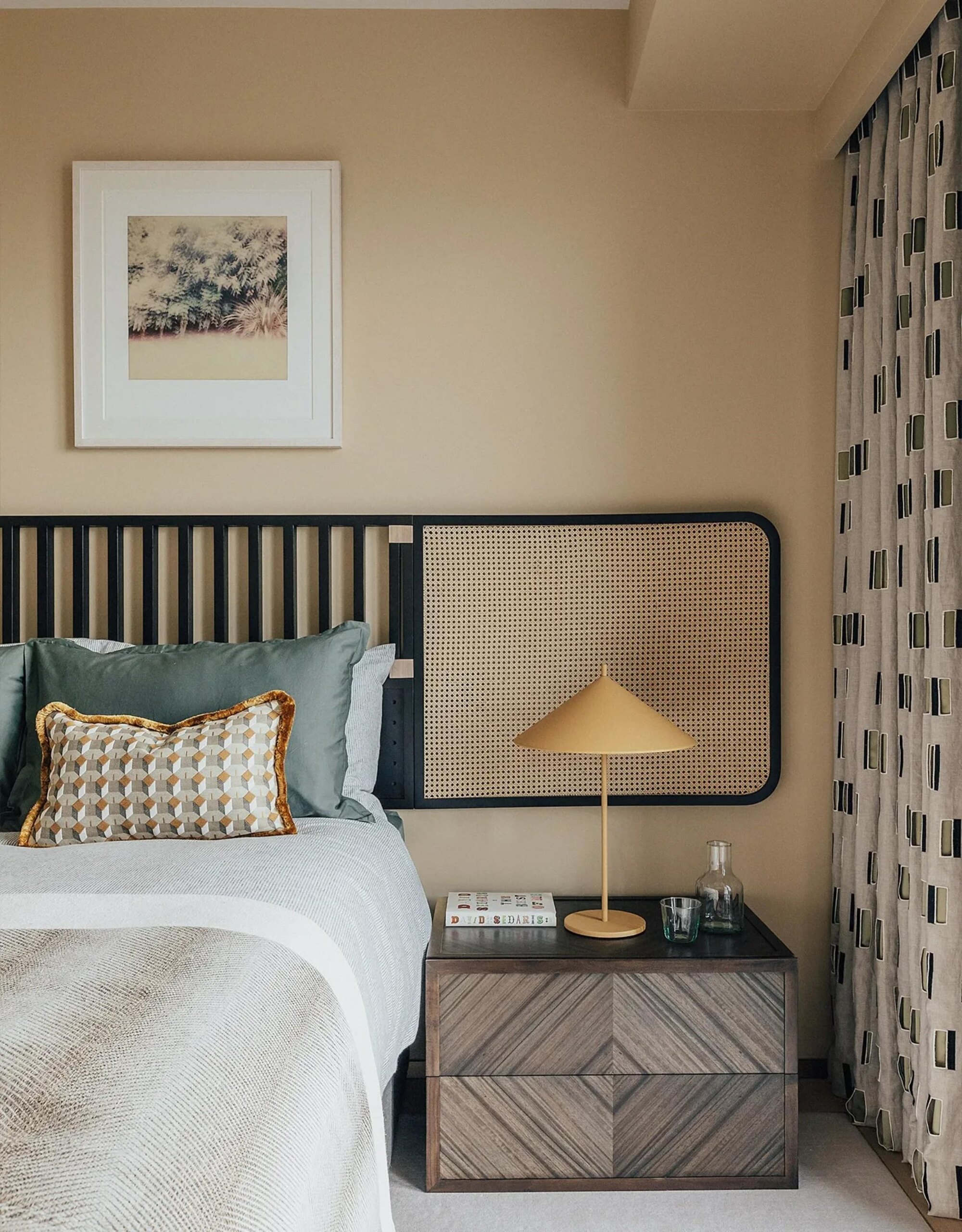beige bedroom with extended wall-mounted headboard, artwork, patterned curtains, timber side table with yellow lamp, and blue cushions