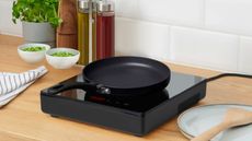 A black single burner electric stovetop on a wooden kitchen counter with a black pan on top. 