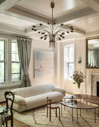 Image of a traditional living room design. There is a white, boucle sofa on top of a white rug with a diamond pattern. In front of the sofa is a wooden pinwheel coffee table. There is a chandelier hanging from the ceiling and a fireplace with a mirror over it in the room.