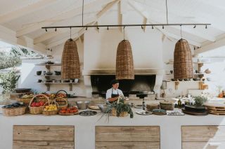 A woman chef is captured at work in an organically furnished show kitchen featuring rattan pendant lights, baskets filled of vegetables, and greenery embracing either one of its sides.