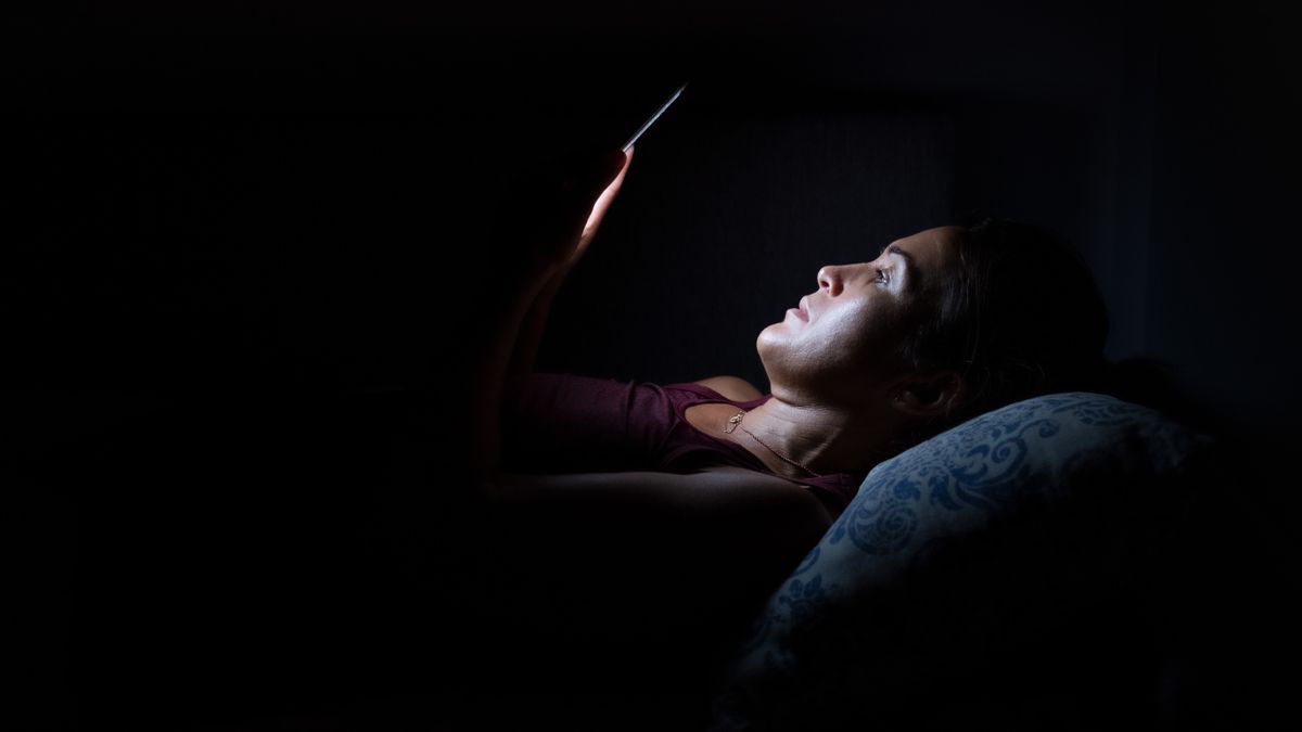 A woman lying in the dark, her face illuminated by the light from her phone screen.