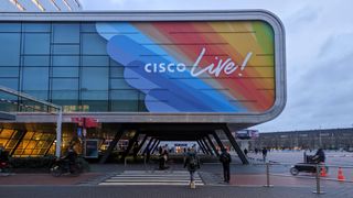 The Cisco Live 2024 sign on the outside of the RAI conference center in Amsterdam, against a background of rainbow colors. Below, cyclists move through the street.