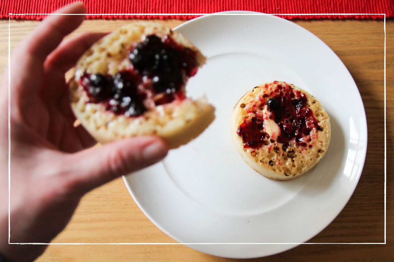 Person taking a bite out of a crumpet with butter and jam on it, while a second sits on the plate