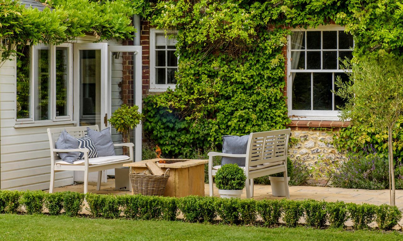 Two wooden chairs with blue patterned cushions next square outdoor fire place next to wall with creeper plants on