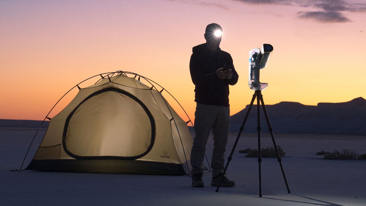 A man with a lit headtorch looking at a smartphone and stood in between a tent and the Vaonis Vespera II - X edition with a sunset in the background.