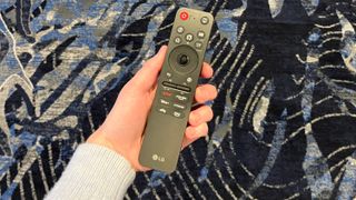 An outstretched hand holding the 2025 LG AI Magic Remote in front of a colorful carpet