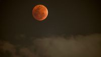 A red full moon is pictured above grey clouds in a dark night sky.