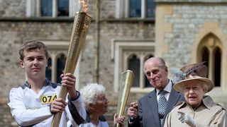 Queen Elizabeth II and Prince Philip, Duke of Edinburgh look on as Olympic torch bearer Gina Macgregor hands the flame to Phil Wells at Windsor Castle on day 53 of the London 2012 Olympic Torch Relay on July 10, 2012 in Windsor, England