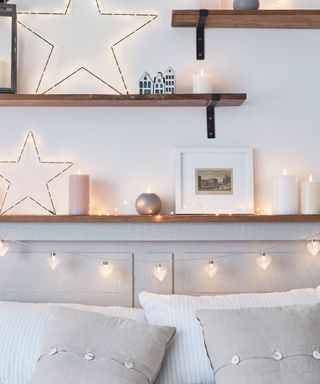 A white and gray bed decorated with gray pillows, with wooden shelves, fairy lights, and star lights above it