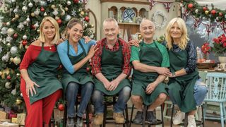 Claire Sweeney, Miquita Oliver, Terry Christian, Sir Tony Robinson and Gaby Roslin wearing green aprons sit in front of a Christmas tree in the tent for The Great British Bake Off Christmas Special.