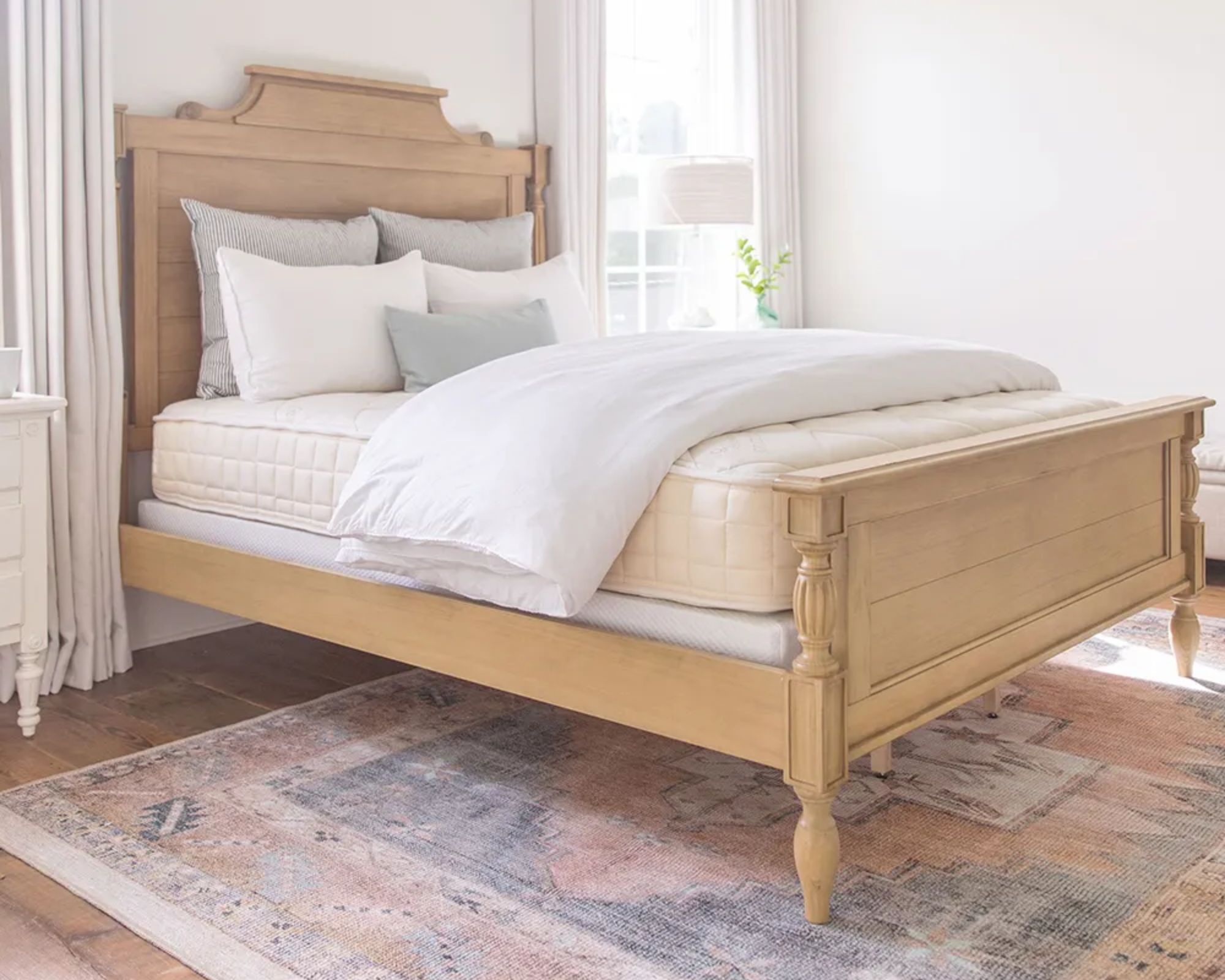 A Naturepedic Chorus Mattress on a wooden bedframe with grey and white cushions, and a folded white duvet, in a bedroom with white walls. To either side of the bed are large windows with white curtains, the flooring is wooden, and underneath the bed is a large Persian-style rug.