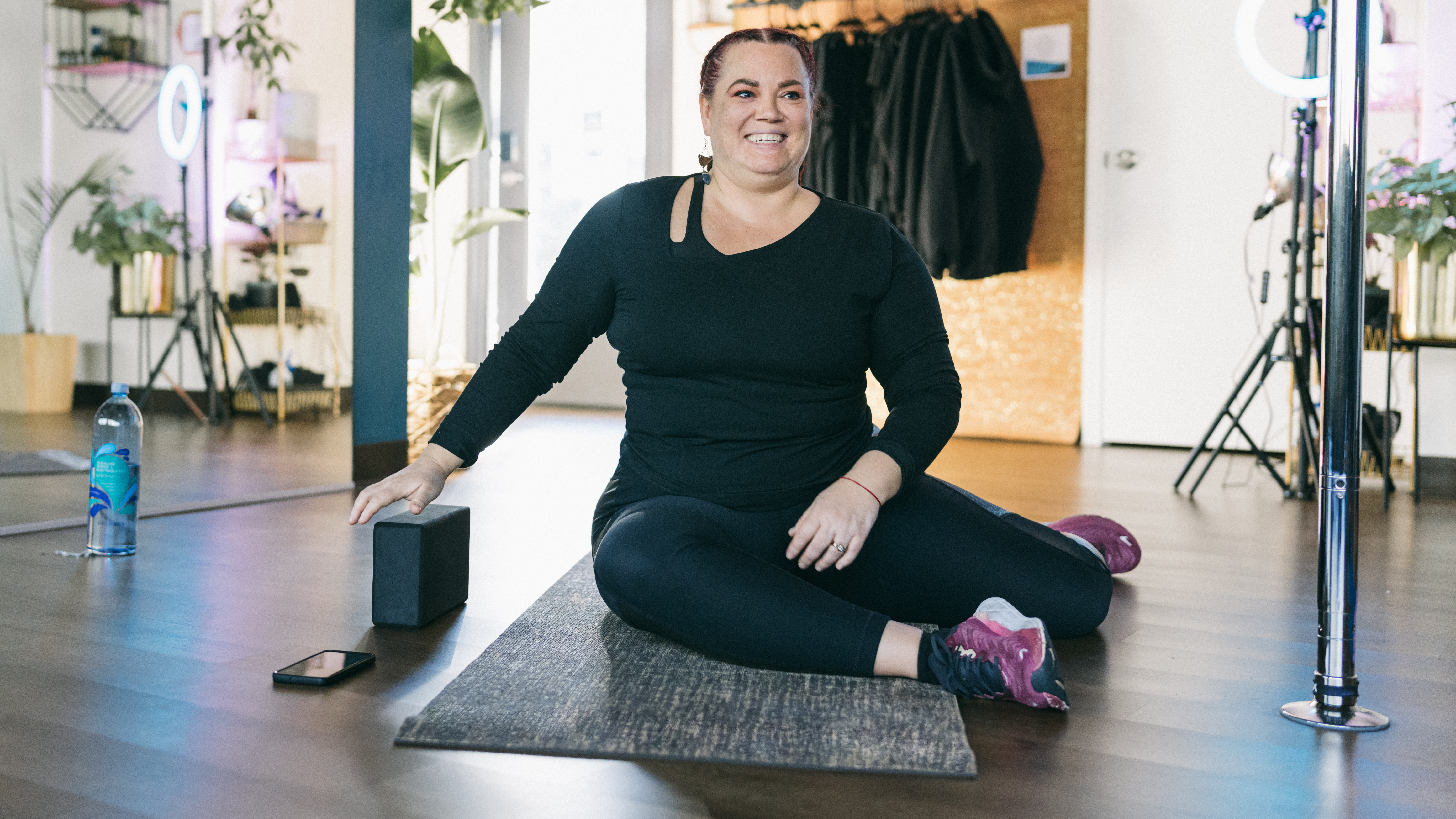A woman in a long sleeved top, leggings and sneakers sits on an exercise mat with her legs bent to her left hand side. There is a yoga block and water bottle next to her. 