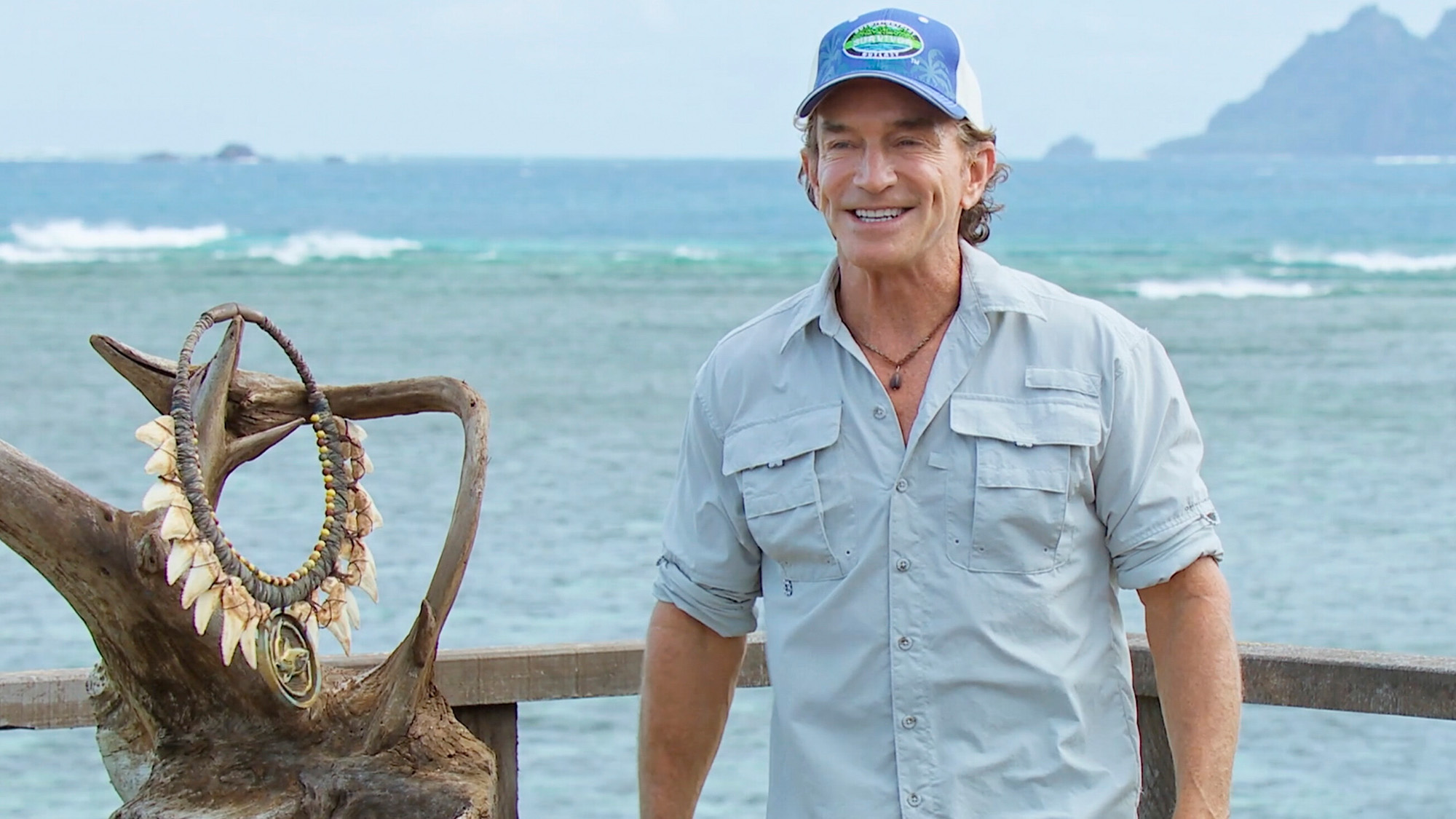 Photo of Jeff in a blue shirt and blue hat announcing a challenge on Survivor. The necklace is in the background.