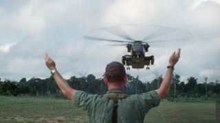 A man stands with his arms to the air as a helicopter approaches Jonestown