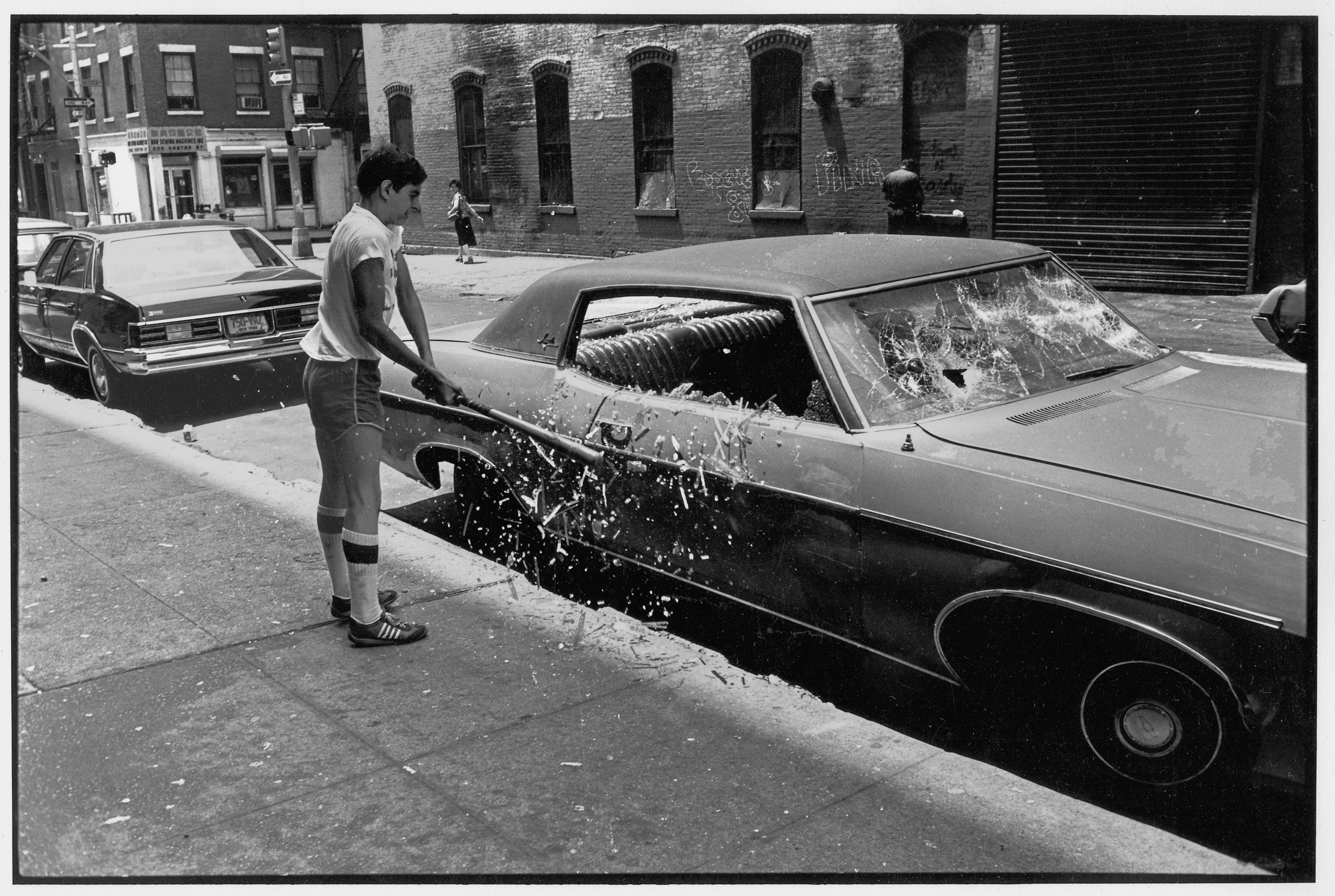 boy in shorts and tube socks swings a baseball bat at a wrecked car, sending shattered glass flying on a New York street