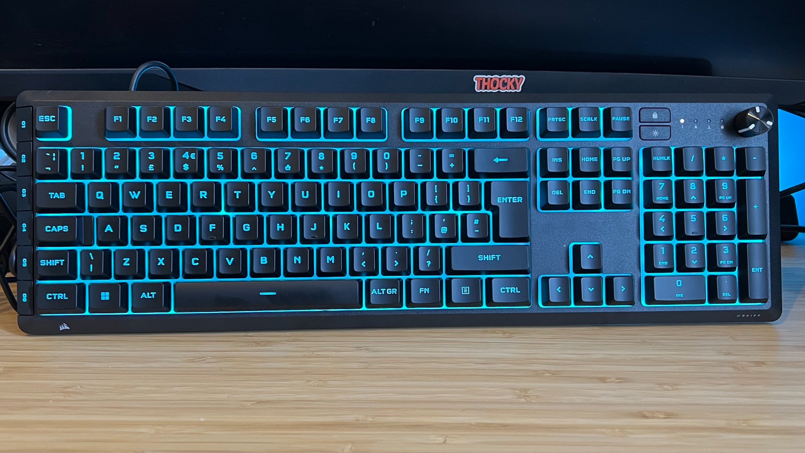 Corsair Skiff 100 Plus gaming keyboard standing upright with blue backlighting on a wooden desk