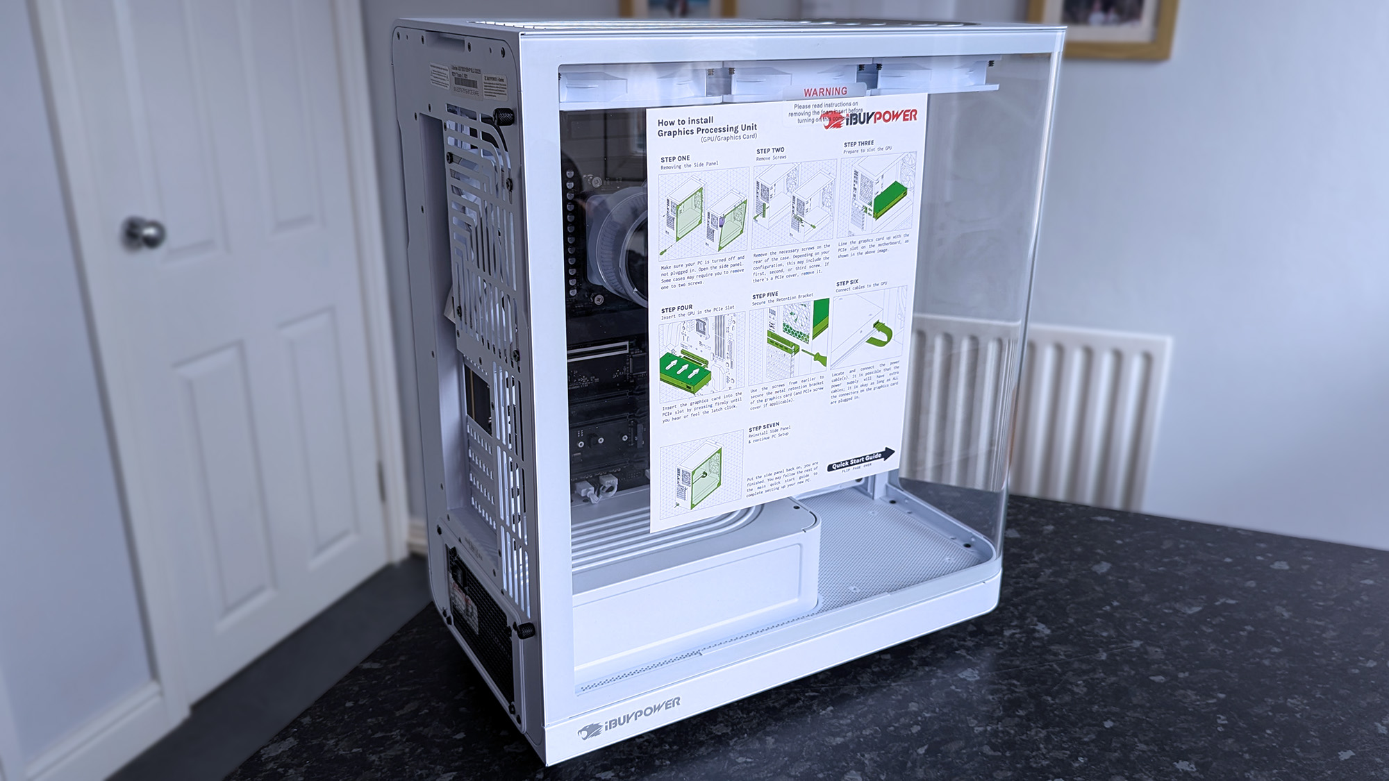 A white iBUYPOWER RDY Trace X R01 pre-built gaming PC sits on a black countertop, its transparent side panel showing a paper with graphics card installation instructions.