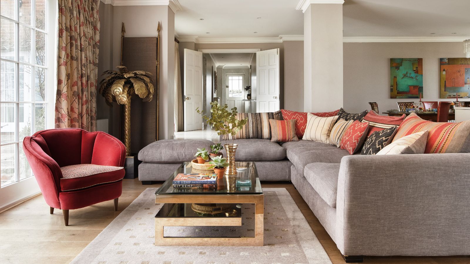 A modern living room with gray L-shaped couch, cream rug, wooden and glass coffee table, red velvet chair and gold statement palm tree in the corner.