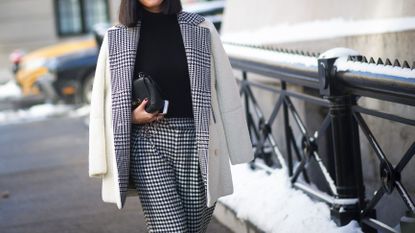 Woman wearing white coat with black and white check lining and black and white check pants holding black purse