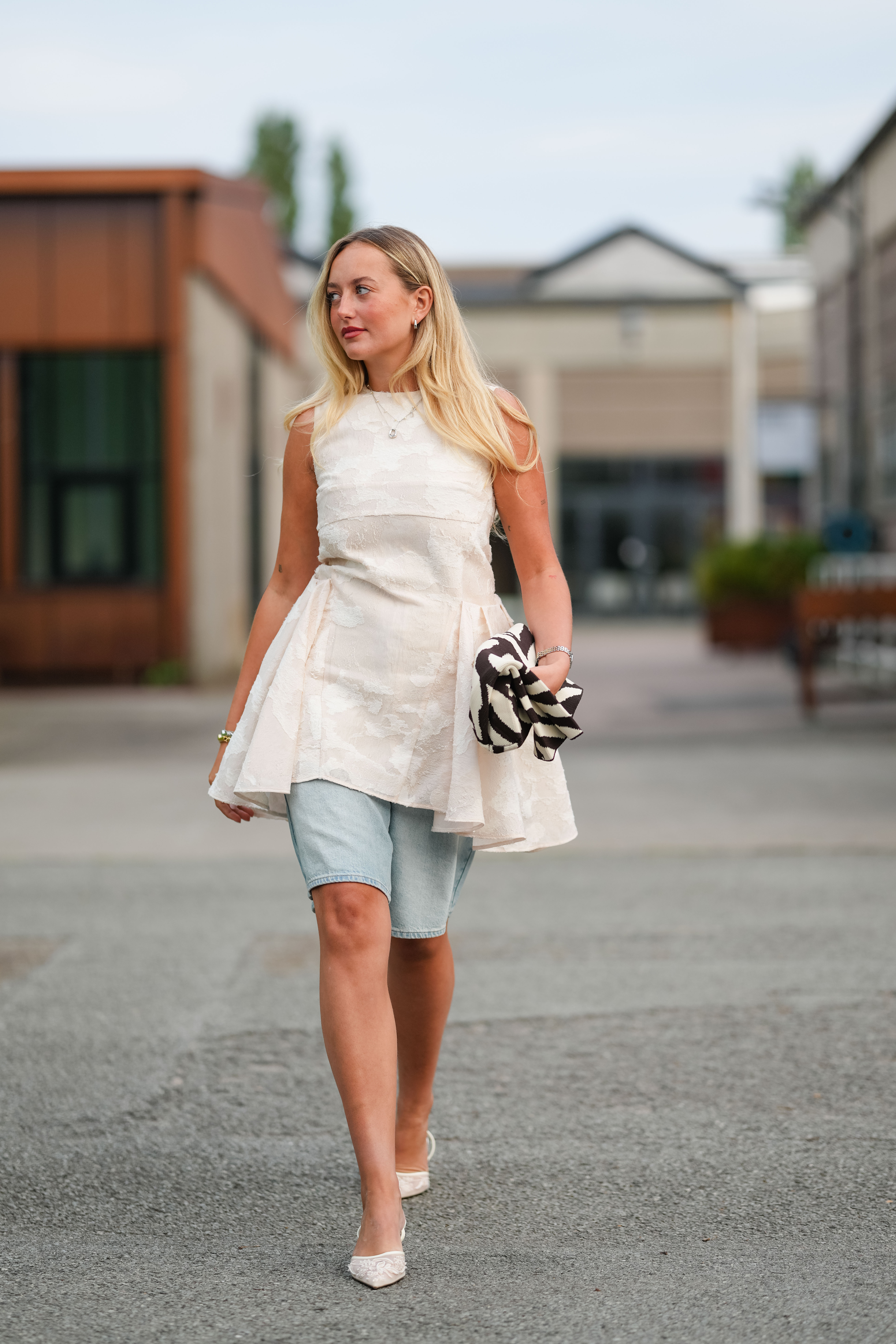 A guest wears long straight blonde hair worn loose with a subtle side part, pink lipstick and light eyeliner, small hoop earrings, a delicate pendant necklace, and gold bracelets with rings; an ivory tonal floral jacquard sleeveless top with a high crew neck, fitted bodice, and dramatic asymmetric flared peplum hem; light-wash denim Bermuda shorts with a finished hem; and white mesh pointed-toe pumps with slender heels, while a pair of black and white striped strappy heels is carried in one hand, walking, outside Rotate, during Copenhagen Fashion Week
