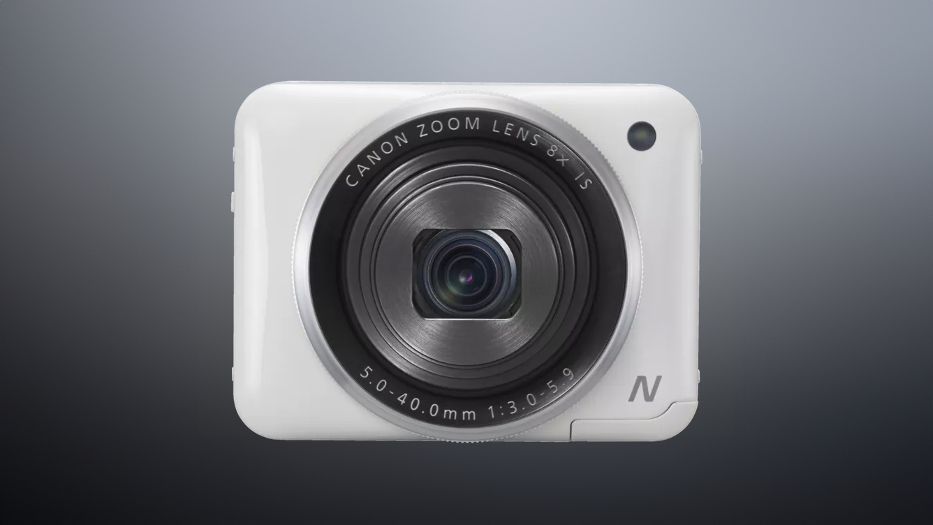 Close-up of a white Canon Powershot N2 compact camera with a zoom lens, featuring specifications on the lens rim, against a gradient background