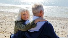 An older couple sits on a beach cuddling, with the woman looking back at the camera, smiling.