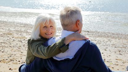 An older couple sits on a beach cuddling, with the woman looking back at the camera, smiling.