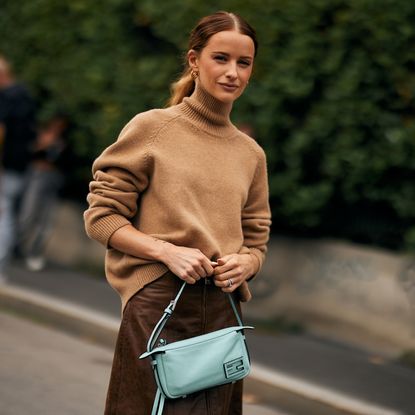 Guest at milan spring 2025 fashion week wearing beige turtleneck sweater and blue handbag 